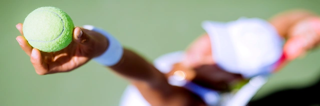 Blurry image showing a tennis player about to serve, holding a tennis ball aloft. The scene conveys a sense of focus and anticipation on a sunny court.