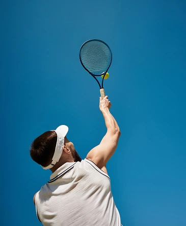 A tennis player, wearing a white visor and white t-shirt, serves under a clear blue sky, concentrating intensely to strike a yellow tennis ball with their racket.
