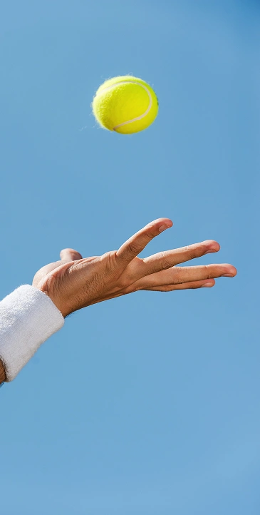 A hand adorned with a white bracelet tosses a yellow tennis ball into a clear blue sky, capturing the motion and anticipation of the serve.