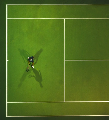 Aerial view of a tennis player preparing to hit a ball on a green court. Shadows and lines create a striking geometric composition.